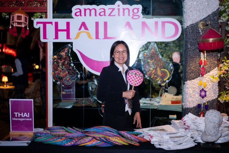Smiling woman in a blazer at a 'Amazing Thailand' booth, holding a pink patterned fan, with colorful textiles on the table behind her.