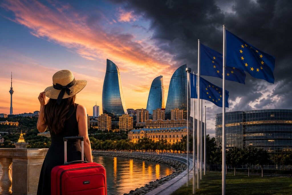 Woman in a black dress and straw hat stands with a red rolling suitcase on the waterfront at sunset, facing the Flame Towers and EU flagpoles.