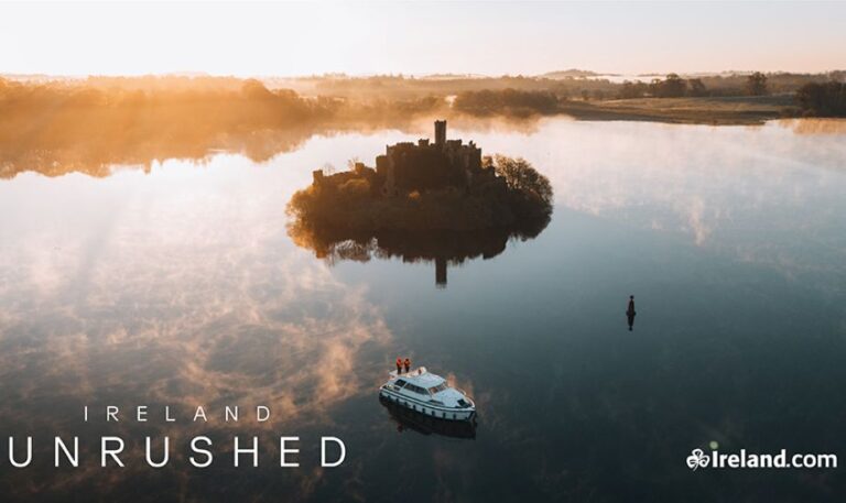 Aerial view of a small island castle in a calm lake at sunrise; a boat drifts nearby (Ireland Unrushed).