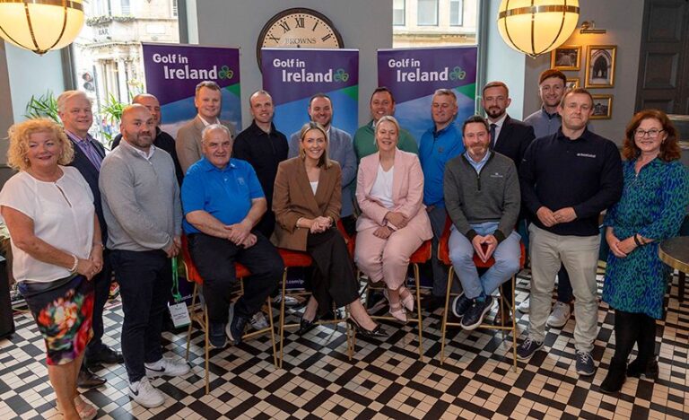 Group of 16 people posing for a photo at a Golf in Ireland event, with banners and a clock in the background in a venue with checkered floor.
