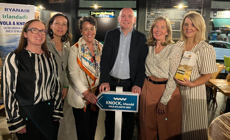 Six adults smiling for a group photo in a café, the man at the center holding a 'KNOCK, Ireland' sign while others hold brochures.