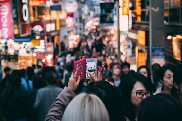 Crowded city street with pedestrians and colorful signs; a person in the foreground takes a photo with a pink phone case.