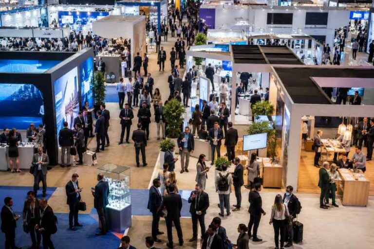 Busy trade show floor with numerous exhibition booths, attendees networking, and wide walkways.