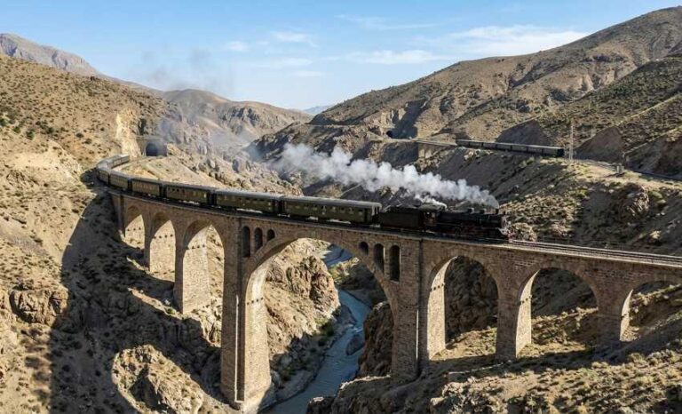 Steam locomotive with passenger cars crosses a tall stone viaduct over a deep canyon and river below, with rugged hills in the background.
