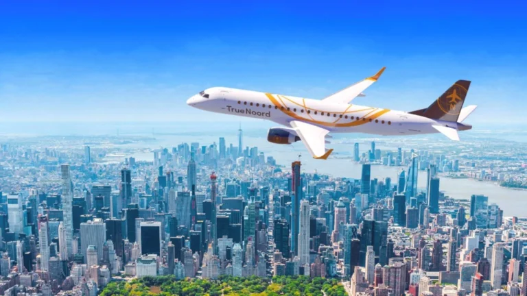White passenger jet with orange accents flying above the Manhattan skyline, TrueNoord logo visible on the fuselage.