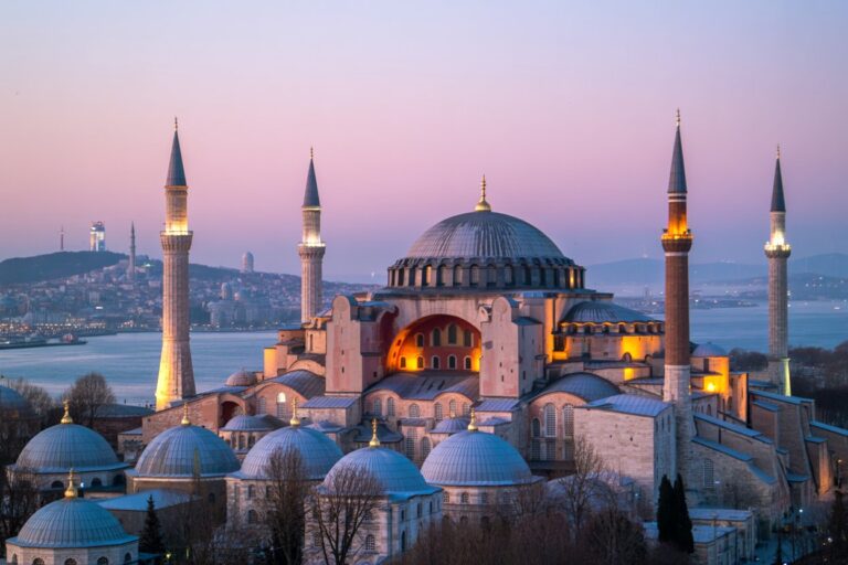 Hagia Sophia in Istanbul illuminated at dusk, showing its large central dome, multiple smaller domes, and four illuminated minarets by the water.