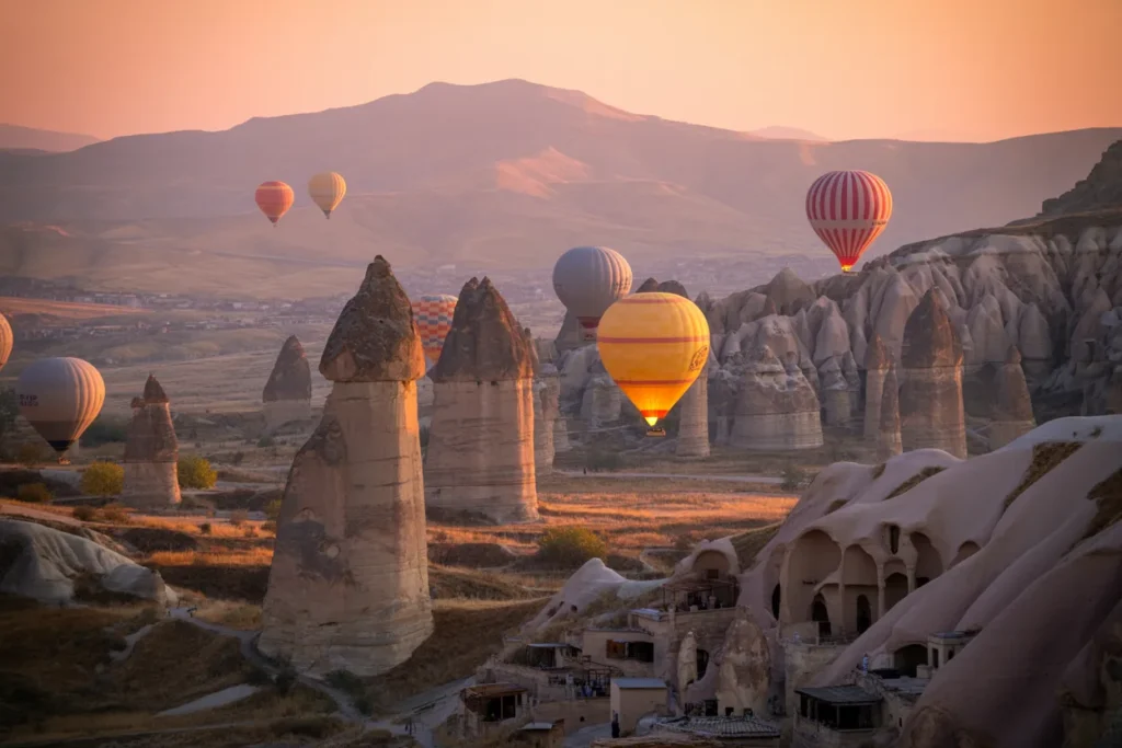Hot air balloons float over tall rock pinnacles and cave dwellings at sunset in Cappadocia, Turkey.
