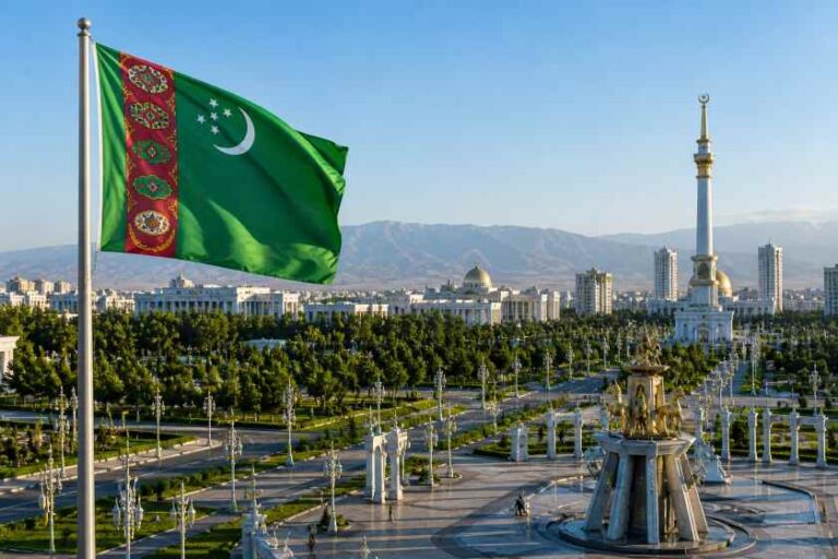 Turkmenistan flag waving over a spacious public square with a fountain, statue, and city skyline in the background in Ashgabat-like setting.