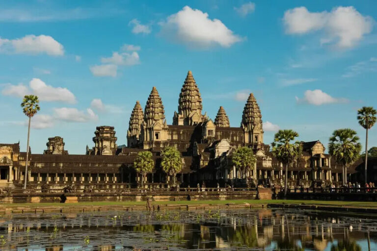 Angkor Wat temple complex with five central towers, palm trees, and a reflective water pool under a blue sky.