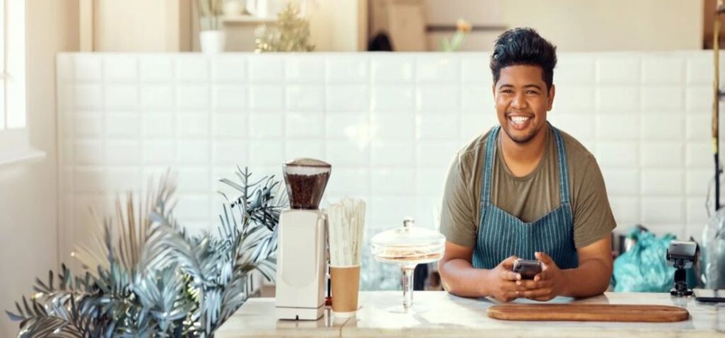 Smiling barista in a blue striped apron sits behind a cafe counter, holding a smartphone with a blender, cups, and cake stand nearby.
