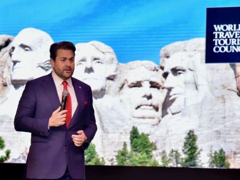 Speaker in a dark purple suit and red tie holding a microphone on a stage with Mount Rushmore backdrop and a World Travel & Tourism Council sign.