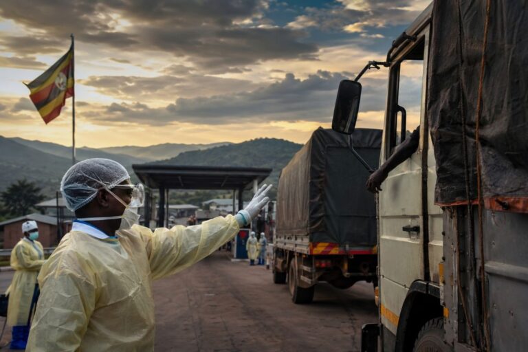 Healthcare worker in full PPE signals to a line of trucks at a checkpoint during sunset, Uganda flag in the background.