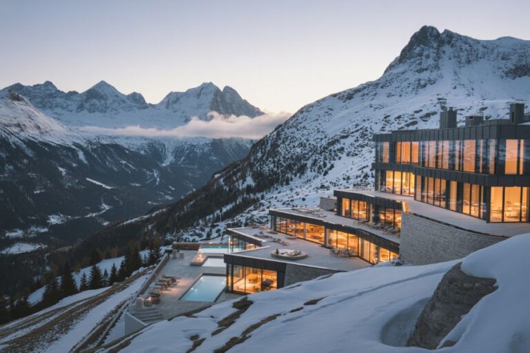 Snowy mountain hotel with glass walls and warm interior lights, perched on a slope at dusk, overlooking a valley and snow-covered peaks.