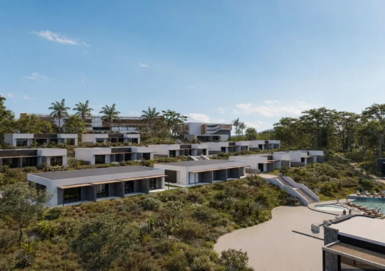 Modern hillside villa complex with white, flat-roofed homes, palm trees, and landscaped terraces surrounding a pool area in the foreground.