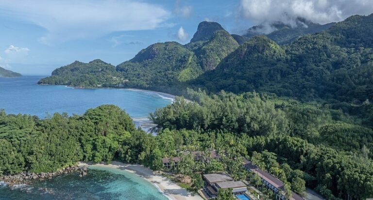 Aerial view of a tropical coastline with a curving white-sand beach, dense green forest, and rugged mountains in the background near a resort complex.