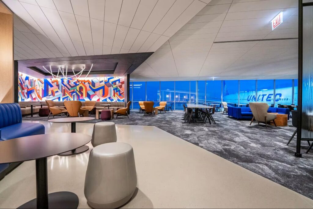 Modern airport lounge with geometric mural, blue couches, and tan chairs around round tables; large glass windows overlook the tarmac.