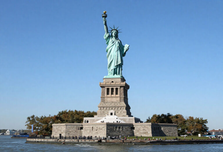 Statue of Liberty standing on a stone pedestal at the harbor, holding a torch and tablet against a blue sky.