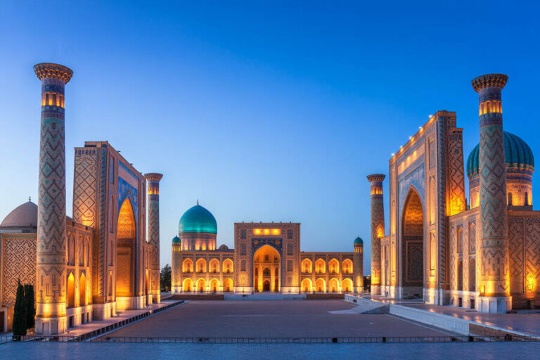 Evening view of an illuminated Islamic architectural complex with turquoise domes, minarets, and a wide courtyard.