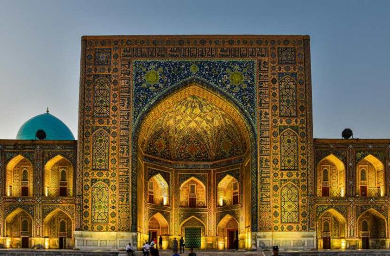 Facade of a richly tiled mosque doorway at dusk, with a blue dome on the left and illuminated arches.