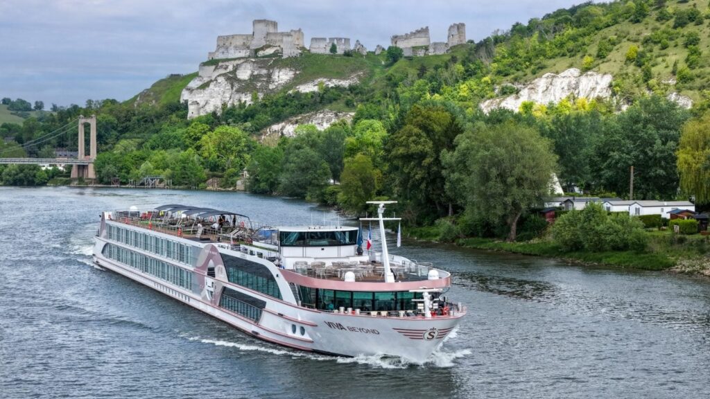 Luxury river cruise ship sailing on a broad river with green hills, a stone castle ruin on a hilltop, and a suspension bridge in the background.