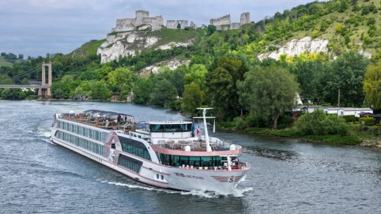 Luxury river cruise ship sailing on a broad river with green hills, a stone castle ruin on a hilltop, and a suspension bridge in the background.