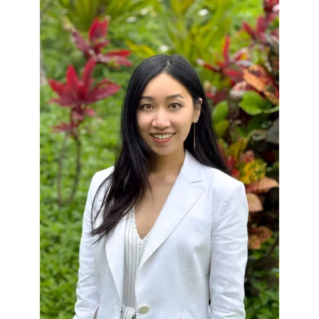 Smiling woman with long black hair wearing a white blazer, standing in a green garden with colorful plants behind her.
