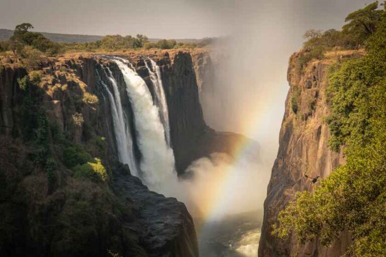 Waterfall plunging between cliffs with rainbow in mist and green vegetation along the rim.