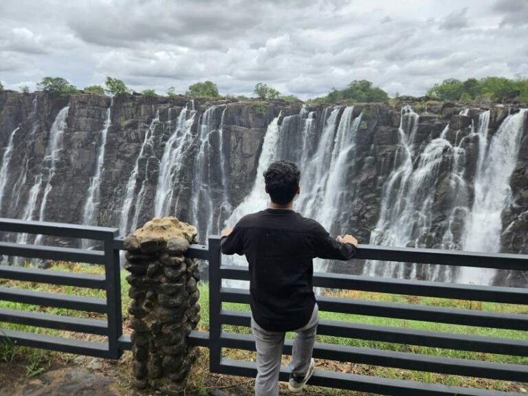 Man standing at a railing, facing away, overlooking a wide cliff with multiple waterfalls cascading down dark rocks under a cloudy sky.
