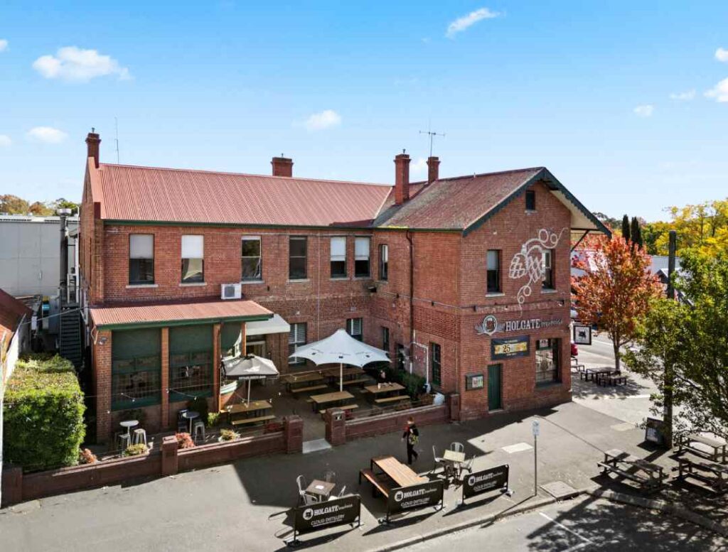Brick two-story building with outdoor wooden tables and benches under umbrellas on a sunny street, Holgate Brewing Co. visible on the facade.
