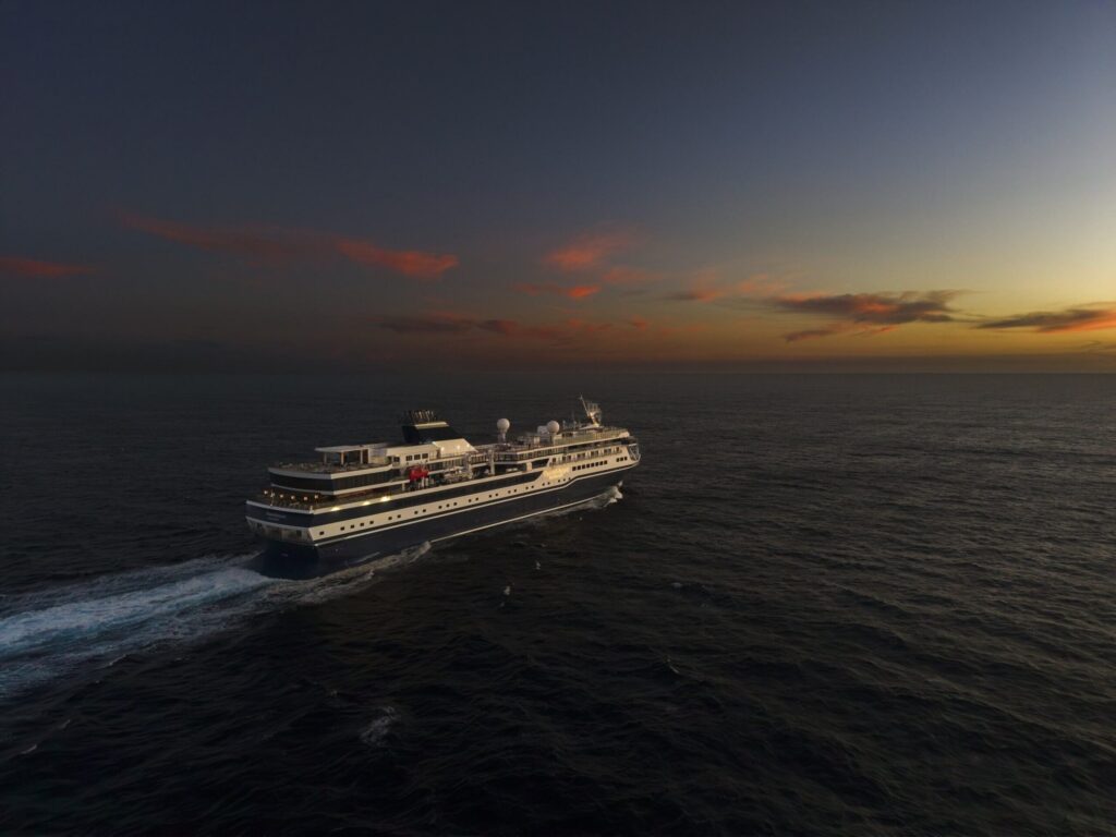 Cruise ship sailing on calm ocean at sunset with orange and pink horizon.