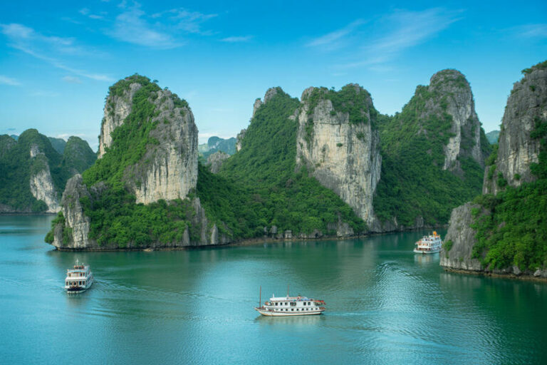 Emerald water with three boats sailing among towering green cliff faces and limestone islets under a blue sky.