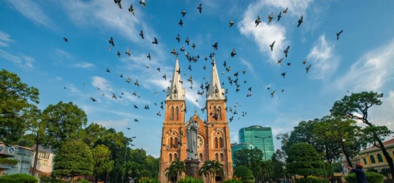 Twin-spired red-brick cathedral with a statue in front, framed by trees as a flock of birds flies overhead against a blue sky.