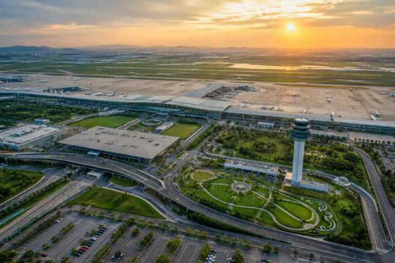 Aerial view of an airport at sunset: runways, terminal buildings, a tall control tower, and surrounding roads with green spaces. Subtle note: describes overall layout and key elements visible in the scene.