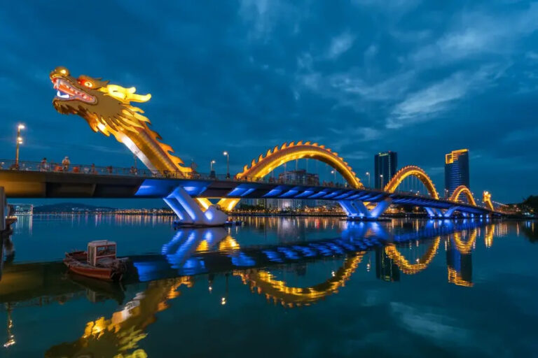 Night view of a dragon-shaped bridge lit with warm yellow lights arching over a calm river, reflections shimmering below.