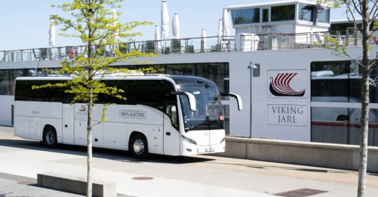 White 100% electric coach bus parked at a Viking Line terminal beside a docked ferry building in the background.