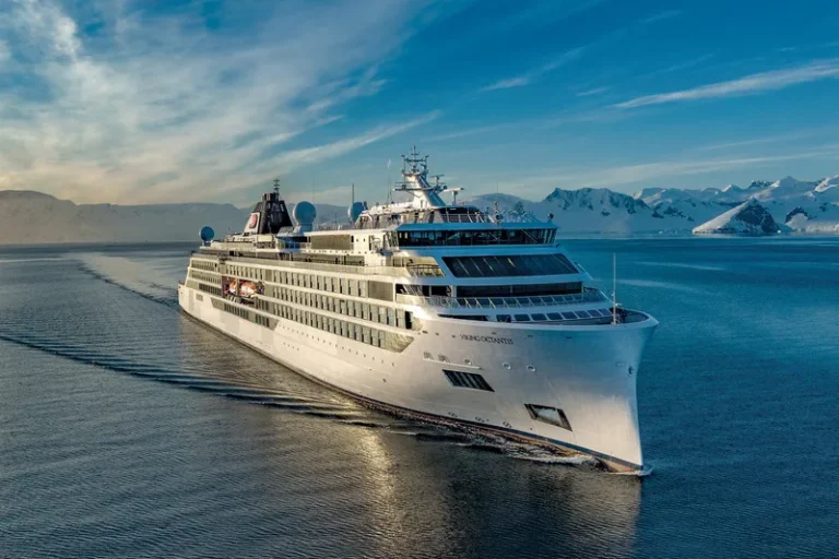 Large white cruise ship sailing through a calm icy fjord with snow-covered mountains in the background.