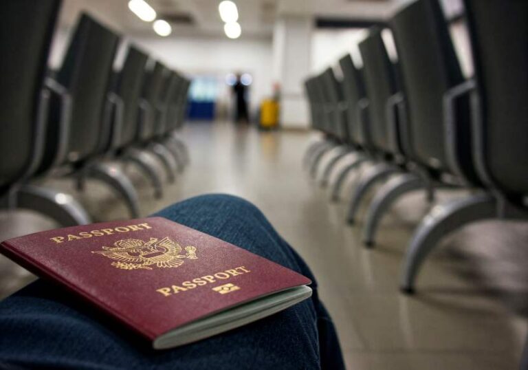 A burgundy passport rests on a person’s knee in a quiet, empty airport terminal seating area, foreground focus.