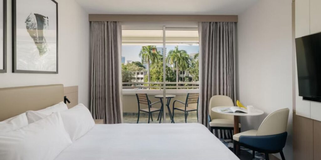 Hotel room with a white bed, sliding balcony doors, and a small table with two chairs on the balcony outside.