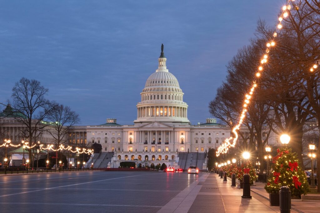 Twilight view of the U.S. Capitol building, illuminated with warm holiday lights along a tree-lined street and decorated Christmas trees on the right.