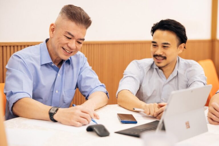 Two men in a meeting: one writes notes, the other looks on at a laptop and phone in a bright office.