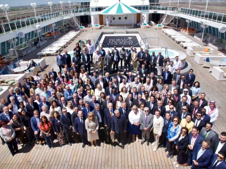 Large group of professionals in suits posing for a group photo on a cruise ship deck, with a pool and lounge area behind them.