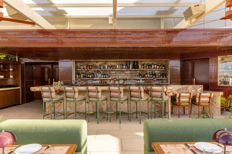 Upscale bar with a marble counter, backlit liquor bottles, and green upholstered bar stools; dining tables in the foreground.