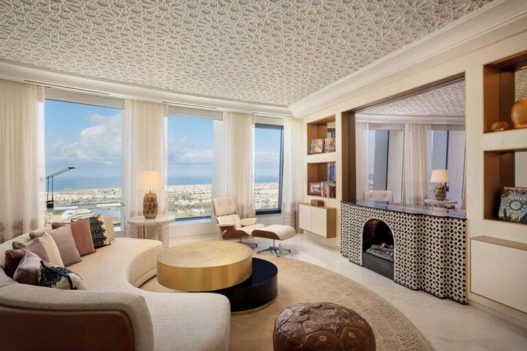 Luxurious living room with a curved beige sofa, gold coffee table, and large windows overlooking a city and sea view at daytime.