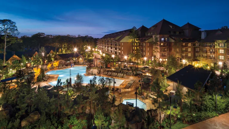 Nighttime view of a resort with a large illuminated pool, lounge chairs, and surrounding trees and buildings glowingly lit along walkways.