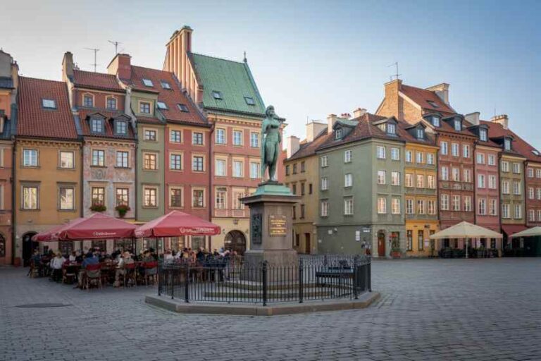 Colorful historic buildings surround a cobblestone square with a statue on a pedestal and outdoor cafe seating under red umbrellas.