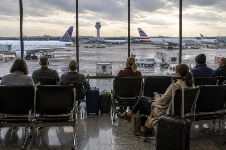 Travelers seated in an airport terminal facing large windows, watching planes on the tarmac outside.