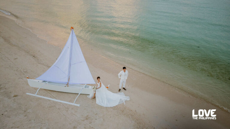 Bride in a white gown and groom in white suit stand on a sandy beach beside a small white sailboat with turquoise water behind them. [Love the Philippines]