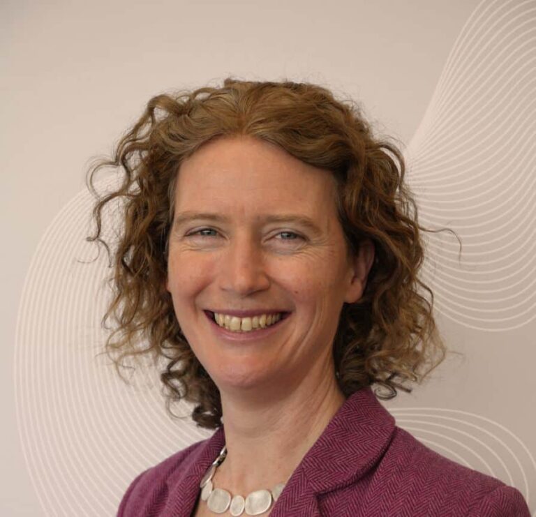 Smiling woman with curly light brown hair wearing a purple blazer, posing for a professional headshot against a beige background with circular line patterns.