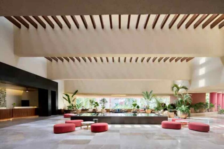 Spacious hotel lobby with red round ottomans and potted plants near a reception desk on the left.