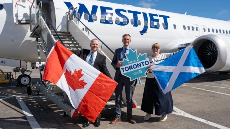 Three staff pose on a tarmac in front of a WestJet aircraft, waving a Canadian flag, a Scottish flag, and a Toronto sign.
